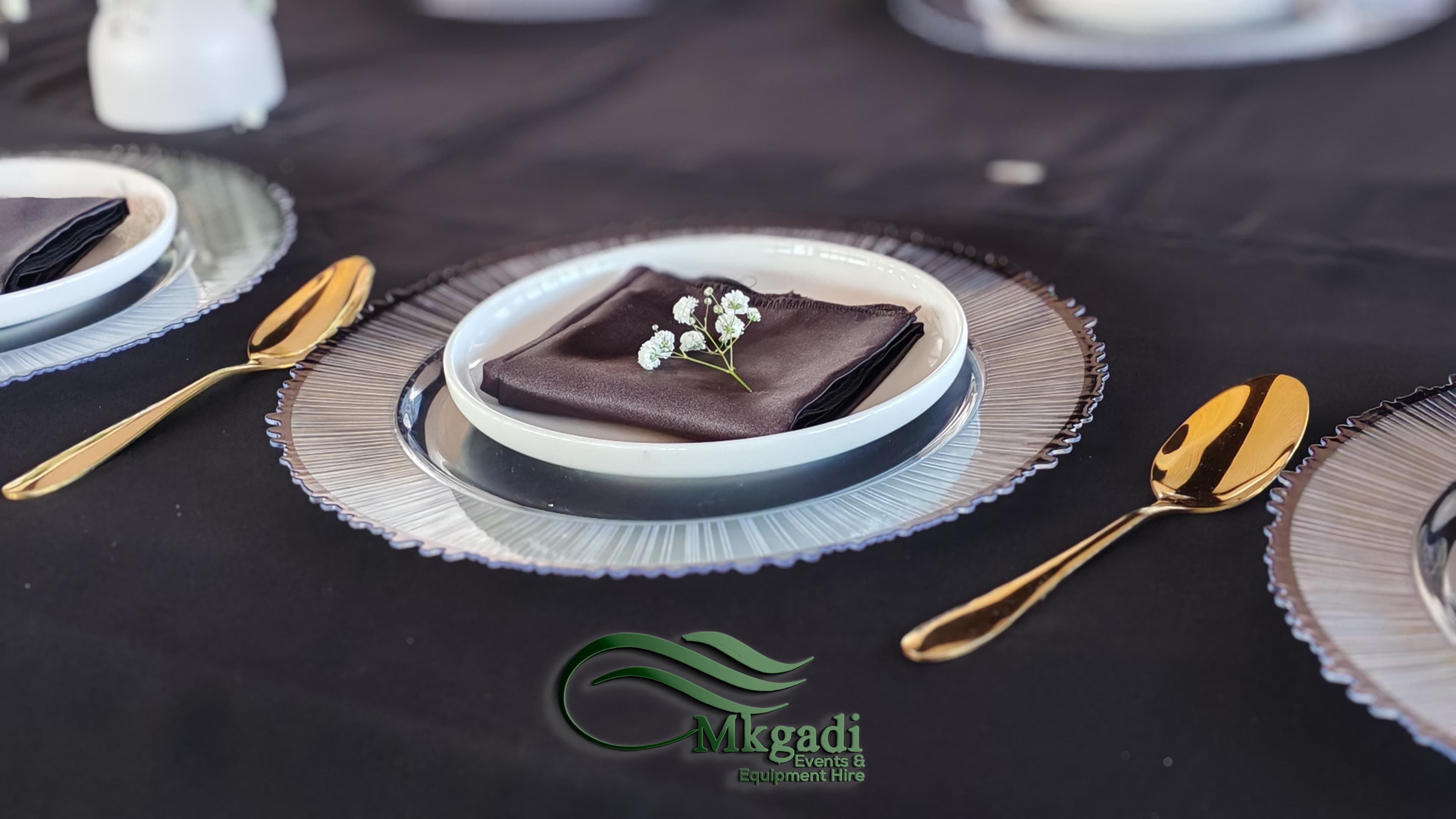 Formal place setting with gold cutlery, glass charger plate and dark linen napkin with white flowers for a dignified occasion