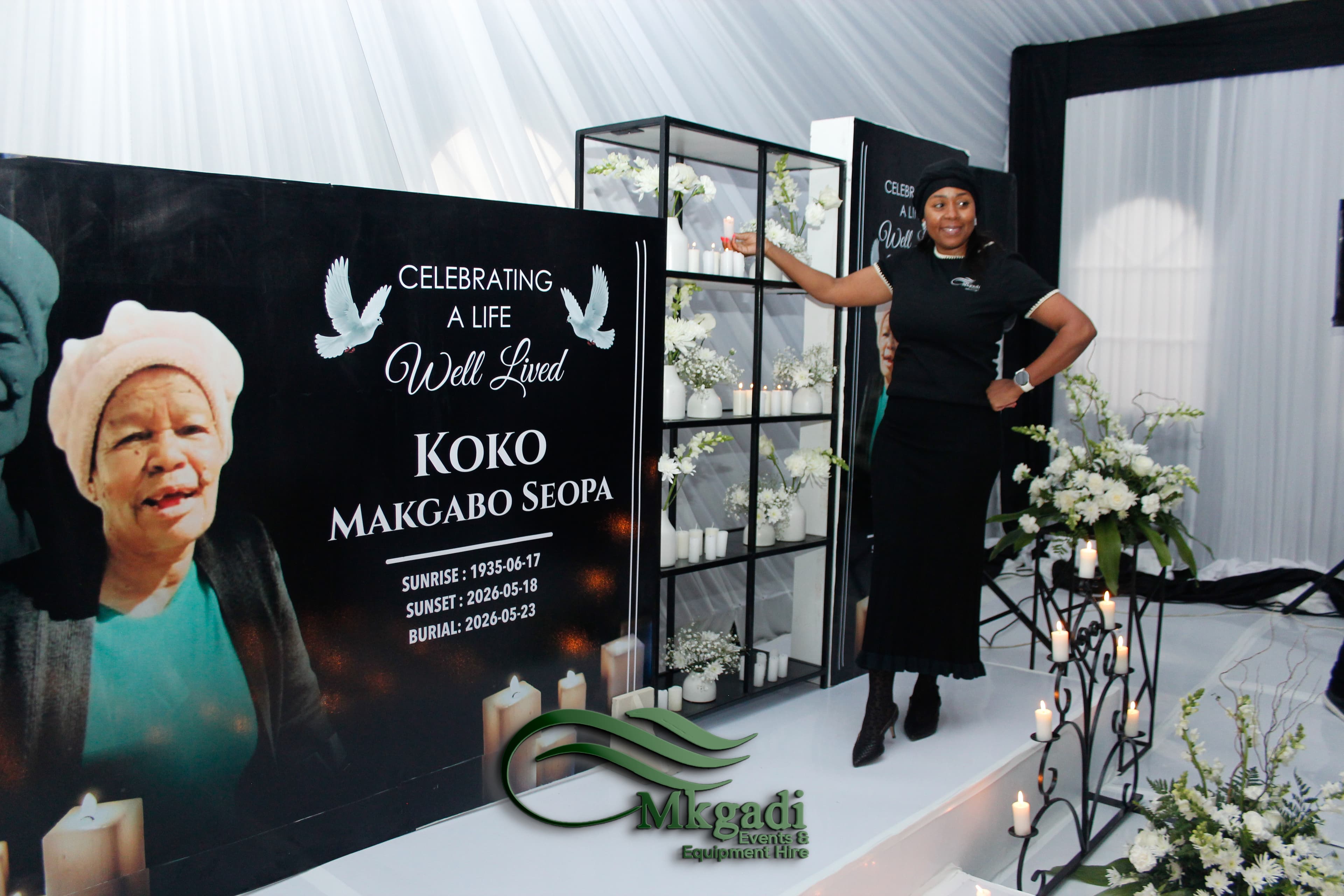Indoor memorial service with a large "Celebrating a Life Well Lived" backdrop, candles, white flowers and a glass display shelf